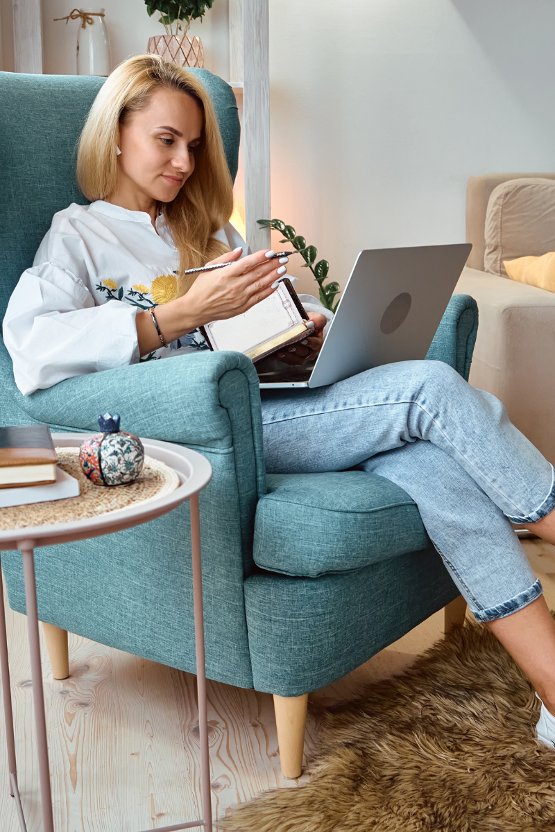 Woman sitting in a blue high-backed armchair with a laptop on her knee, holding a pen and notebook. She is in a room with a sand coloured sofa, and a side table with books and a small decorative pot on it. A small section of rug is also visible.