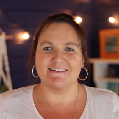 A photo of a white woman (Helen Soutar) with brunette hair.  She is wearing silver hoop earrings and a white v neck top.  A chain is visible around her neck.  She is standing in front of a small set of white shelves with a plant and some textile art on them.  The wall behind her is painted midnight blue and features a string of lights and a macrame wall hanging 