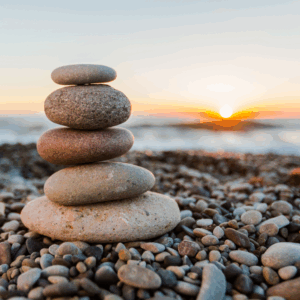 A stack of smooth, rounded stones balanced on a pebbled beach with the sun setting over a calm sea in the background. Symbolizes stability and foundational balance.